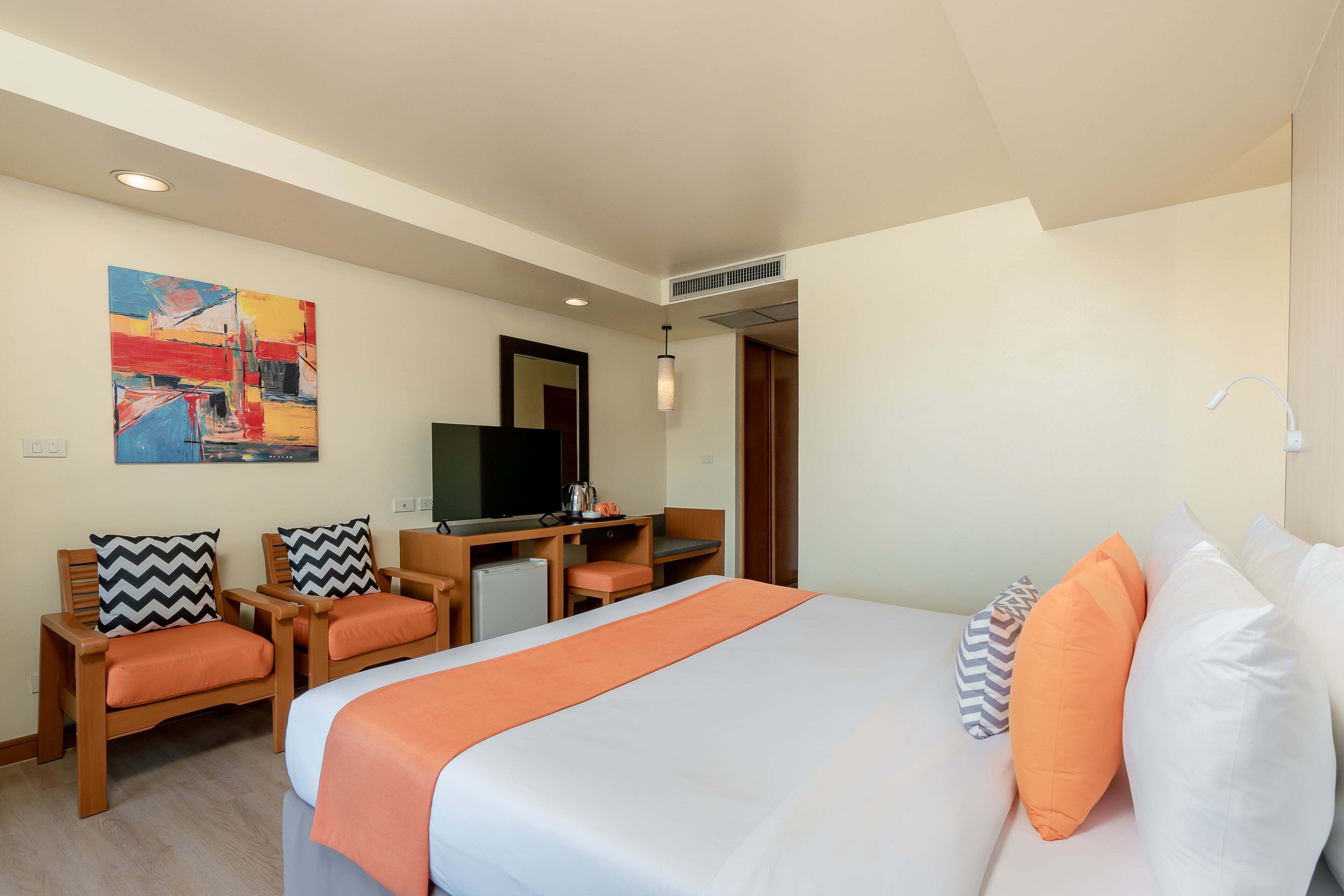 A hotel room with a bed, two chairs, a desk, a television, and a painting on the wall.