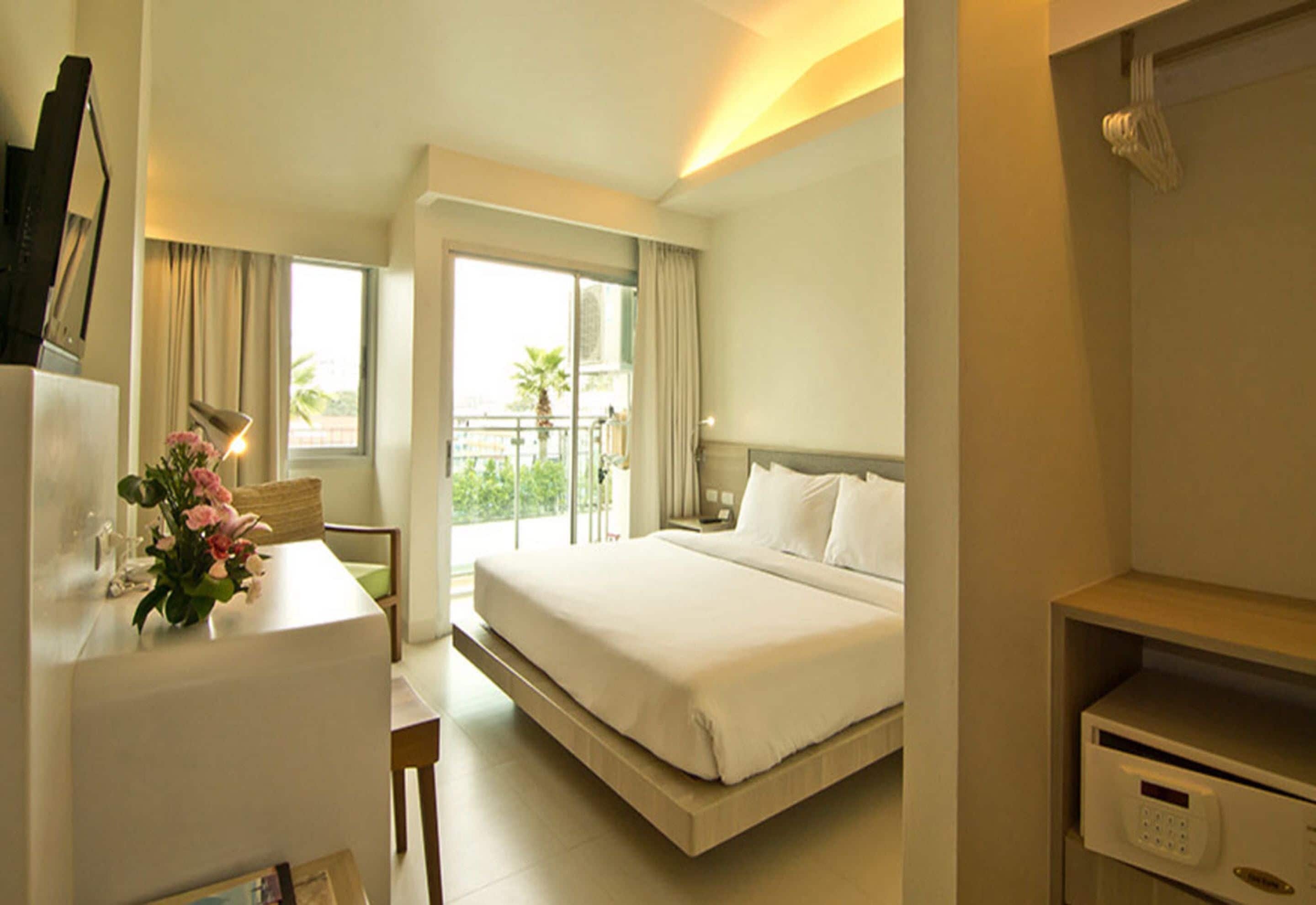 Una camera d'albergo con un letto, una scrivania con una lampada, un tavolino con fiori, un balcone con vista e un pannello di controllo.