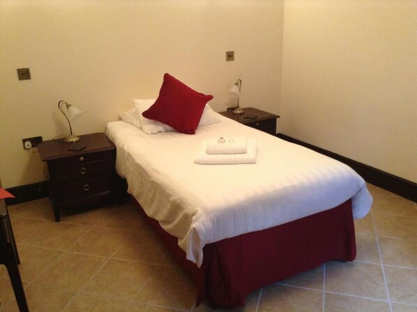 A neatly made bed with a red pillow and white linens, a bedside table with a lamp, and a small drawer unit. - The Crown Hotel (Thetford)