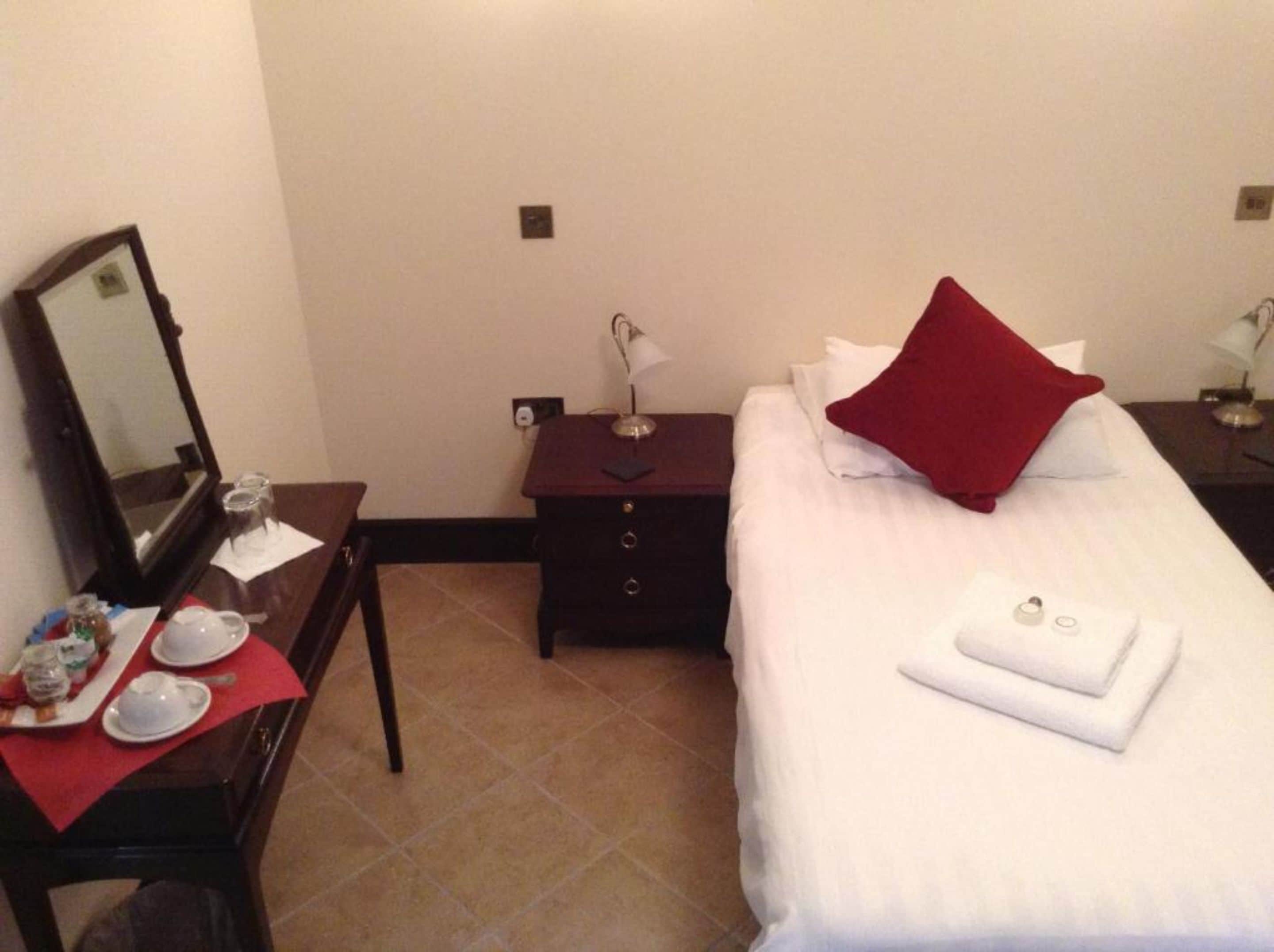 A hotel room with a neatly made bed, a small bedside table with a lamp, a nightstand with a mirror, and a table with a red placemat holding a teapot, cups, and a plate of food.