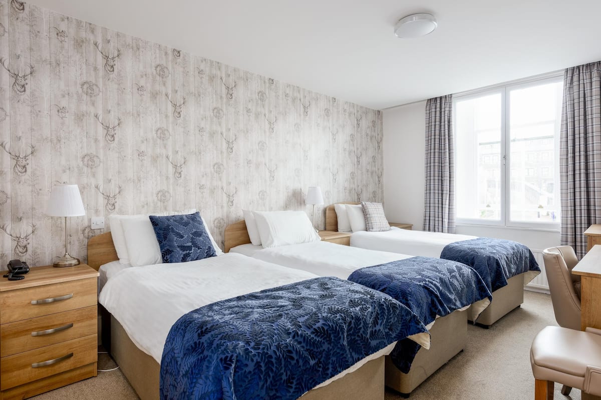 A hotel room with two beds, a wooden dresser, a desk, and a large window with curtains.