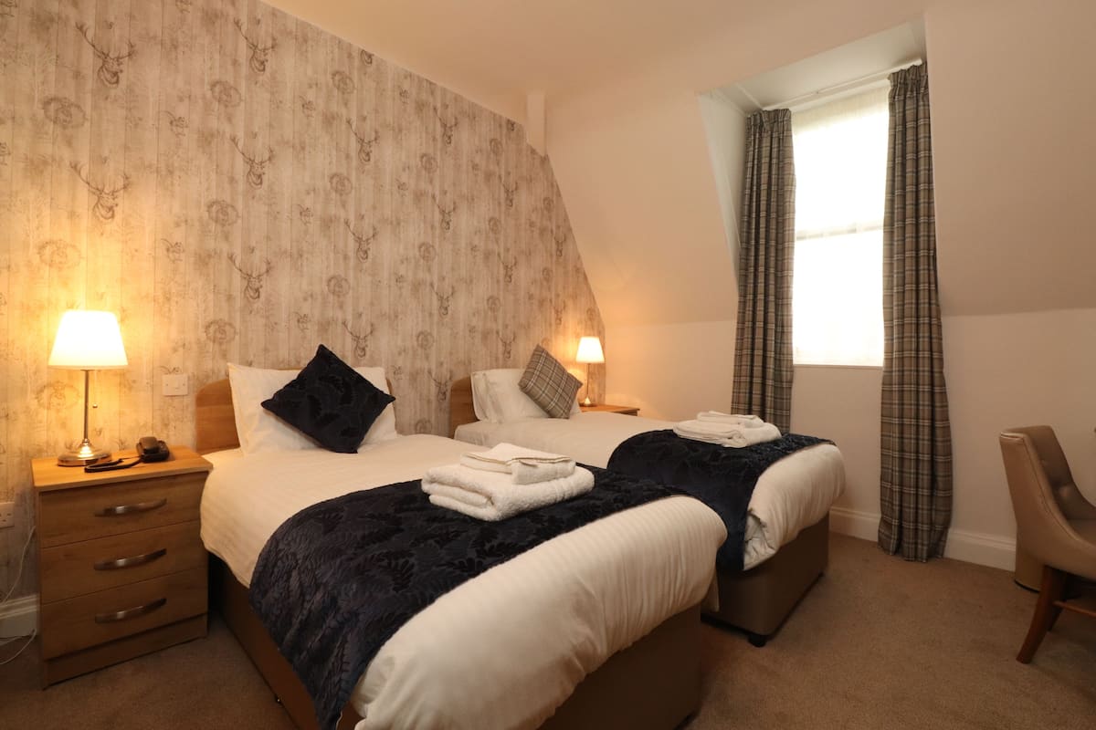A bedroom with two beds, a nightstand with a lamp, a chair, and a window with curtains.