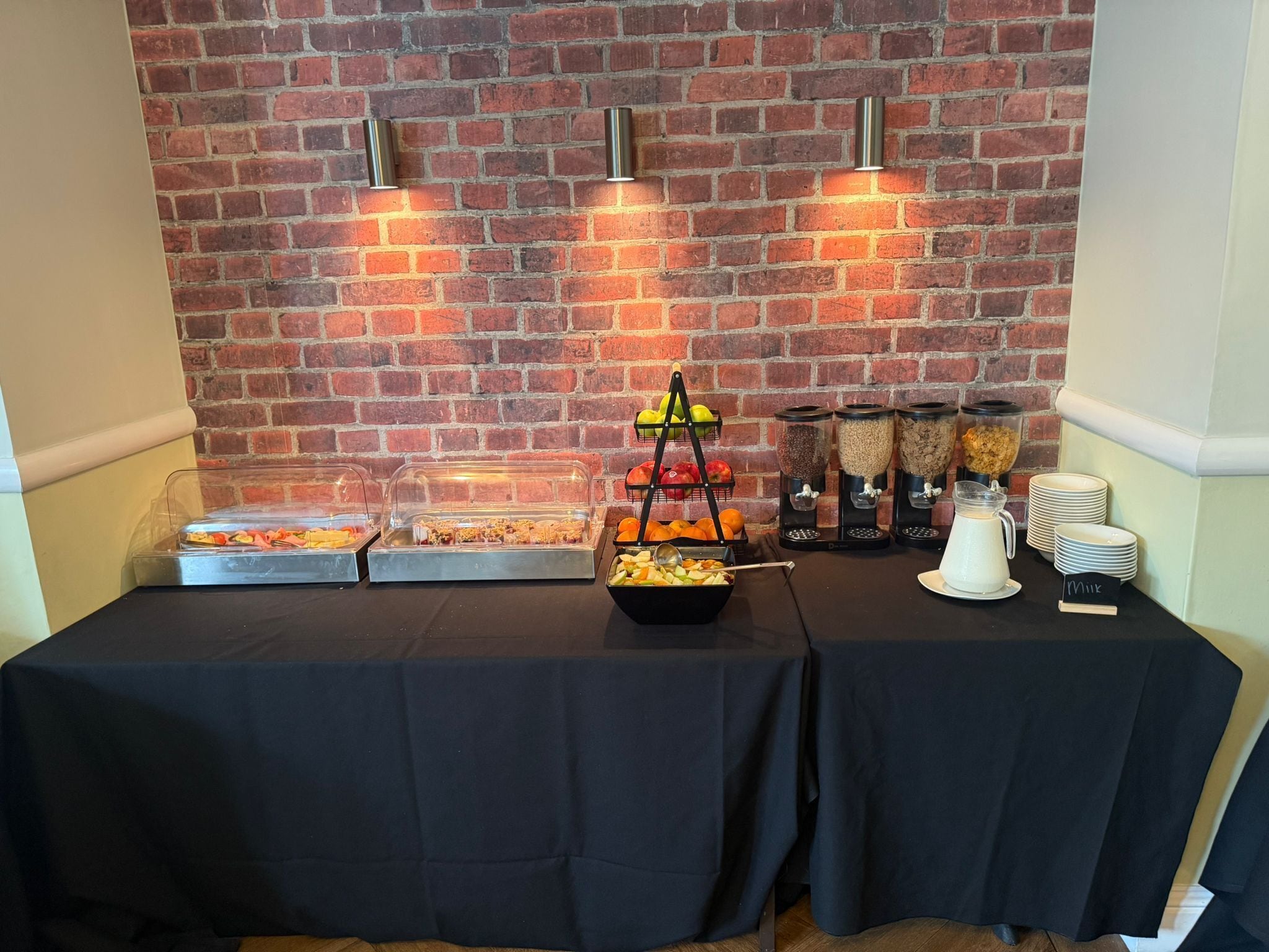 Una mesa de buffet con diversas opciones de comida, entre ellas frutas, cereales y una estación de leche, con un fondo de pared de ladrillo.