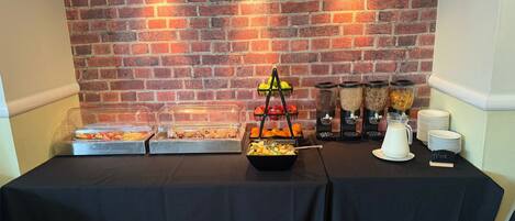 Una mesa de buffet con diversas opciones de comida, entre ellas frutas, cereales y una estación de leche, con un fondo de pared de ladrillo.
