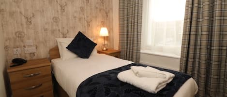 Ein ordentlich bezogenes Bett mit einer dunkelblauen Decke, einem weißen Kissen und einem gefalteten Handtuch auf der Seite. Auf einem hölzernen Nachttisch steht eine Nachttischlampe zusammen mit einem Telefon und einer Schublade. Das Zimmer verfügt über einen Karottenstreifen-Vorhang und ein Fenster mit Blick ins Freie.