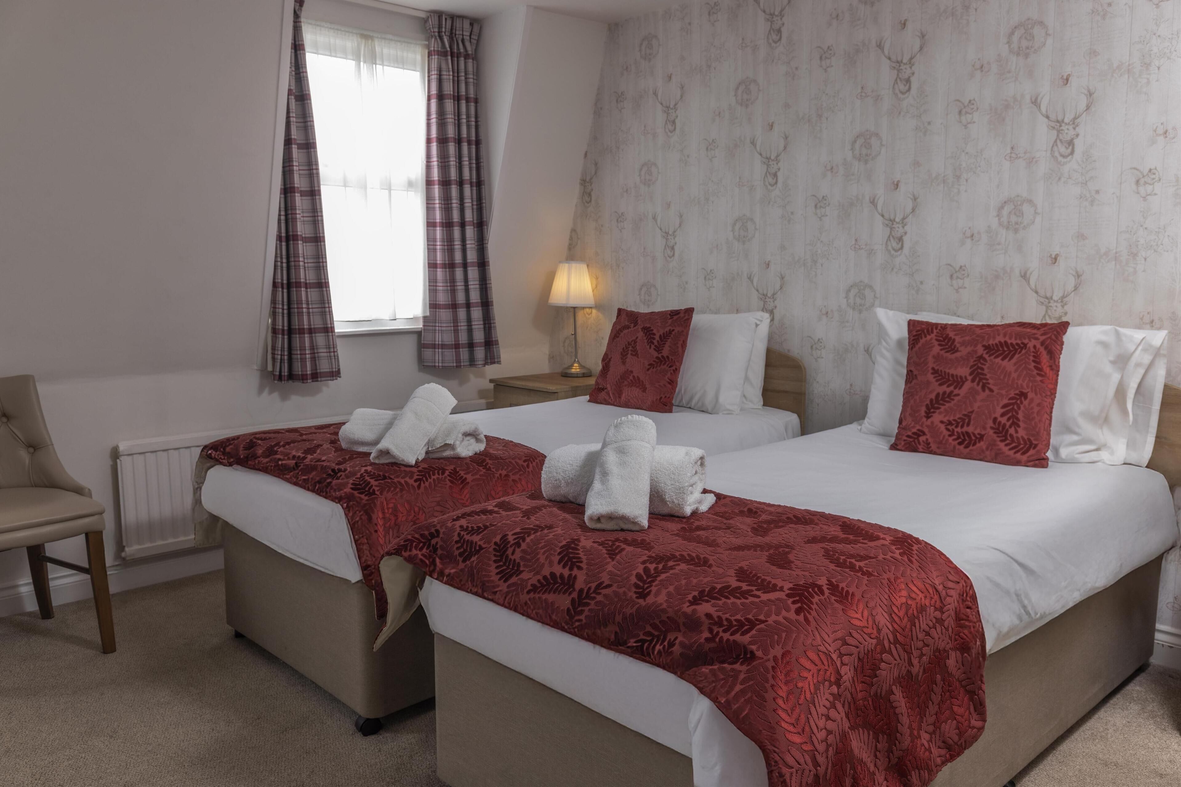 A hotel room with two beds, each with a red blanket and white pillows, a nightstand with a lamp, a chair, and a window with curtains.