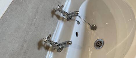 Ein Badezimmer mit einem weißen Waschbecken, zwei Wasserhähnen, einer Holzablage und einer Glasdusche.