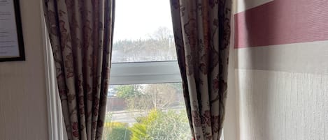 Et værelse med blomstermønstrede gardiner, en radiator, et vindue med udsigt over grøn natur, og et natbord med lampe og lommevæv.