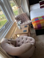 Un dormitorio con una cama, una silla, una mesita con un florero y vistas al exterior.