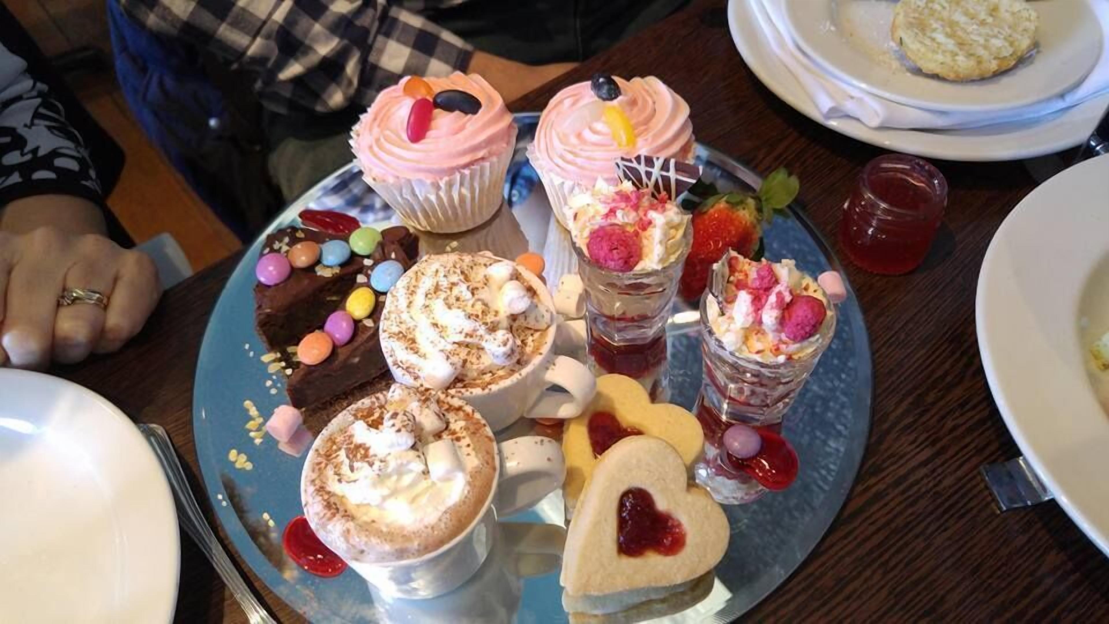 A variety of desserts on a tray, including cupcakes, a heart-shaped cookie, and a mug with whipped cream.