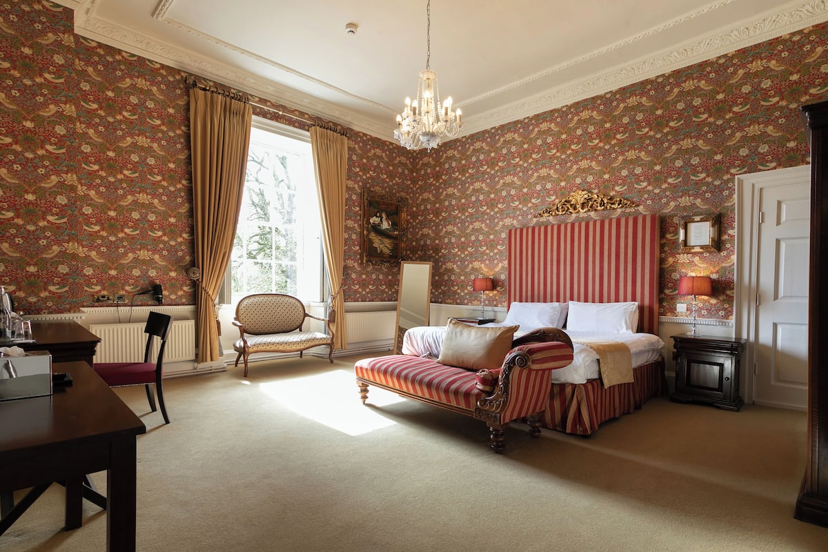 A bedroom with a large bed, a chair, a table, and a chandelier.