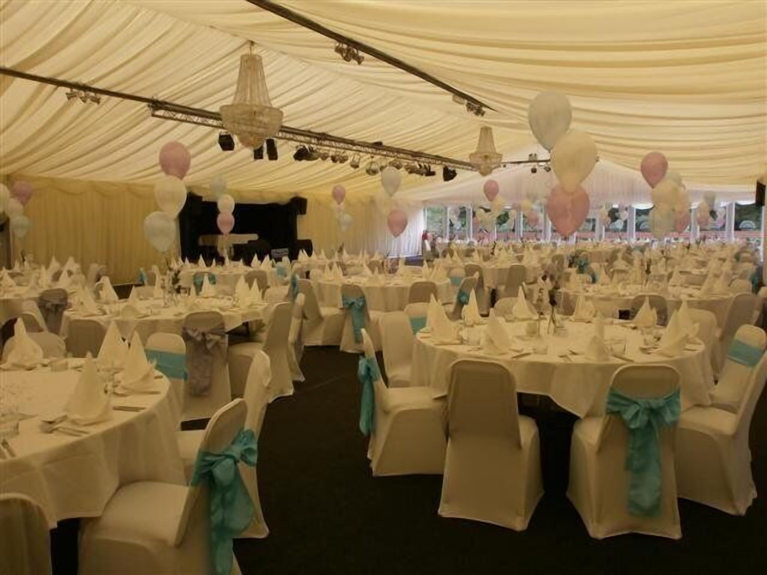 Un salón de banquetes con mesas redondas cubiertas con manteles blancos, sillas con cintas turquesa y un escenario con globos.