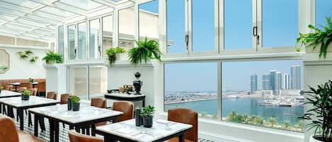 A restaurant with a view of a marina and city skyline, featuring checkered flooring, large windows, and indoor plants.