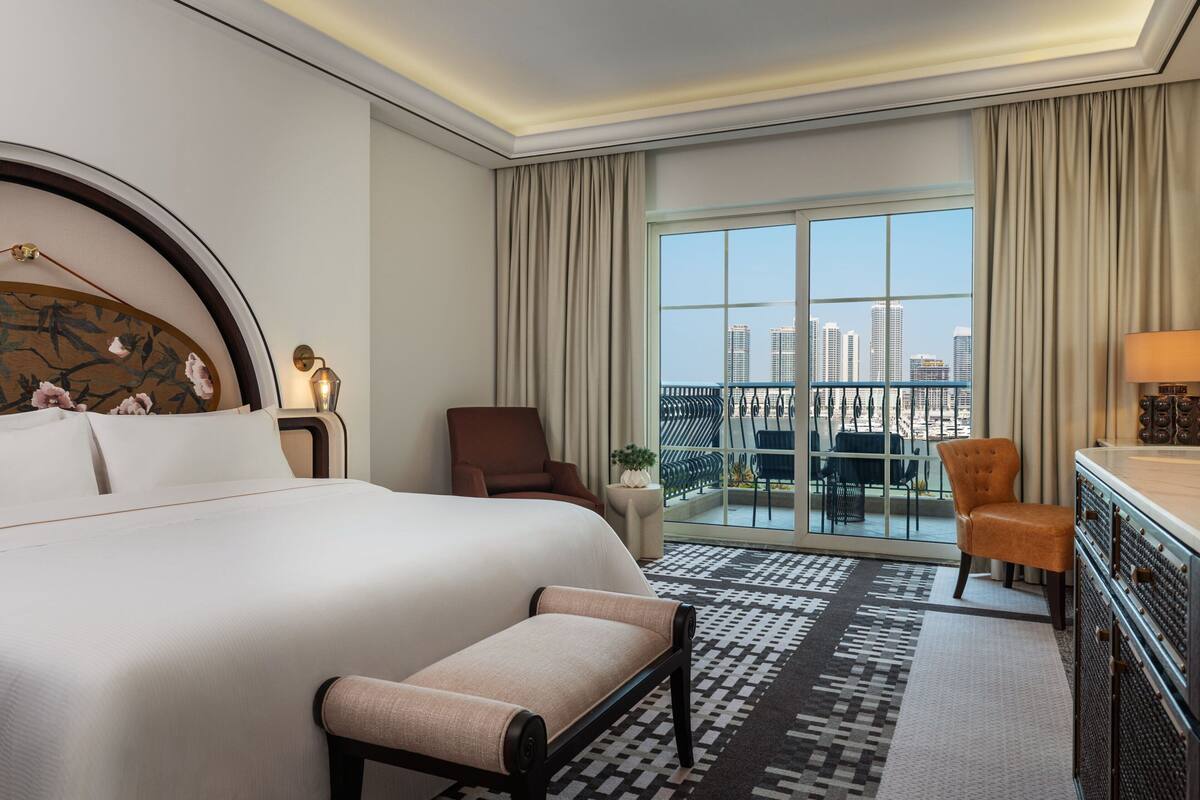 Une chambre d’hôtel avec un grand lit, un bureau, une chaise, un balcon donnant sur la ville et un tapis à motifs.