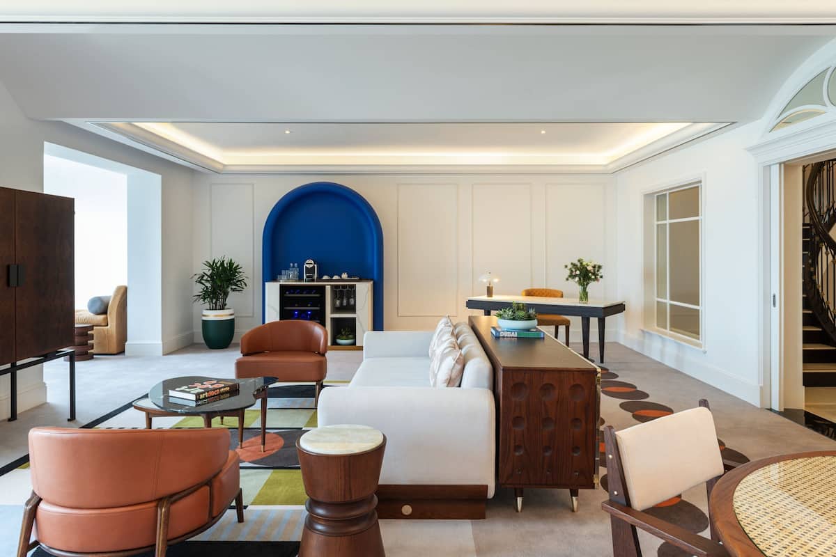 Un grand salon comprenant un canapé, une table basse et des fauteuils. On aperçoit, à l’arrière-plan, une arche bleue et un escalier.