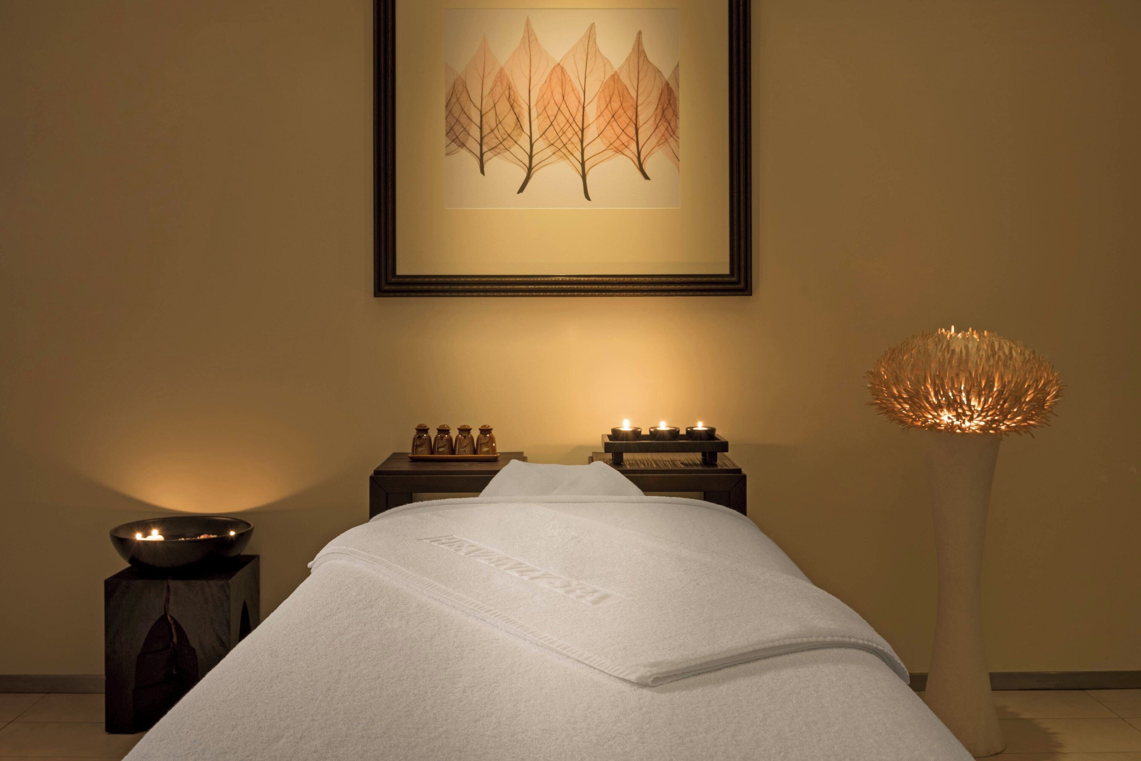 A neatly made bed with a white blanket, a framed artwork of trees on the wall, a small table with candles, and a tall, textured floor lamp.