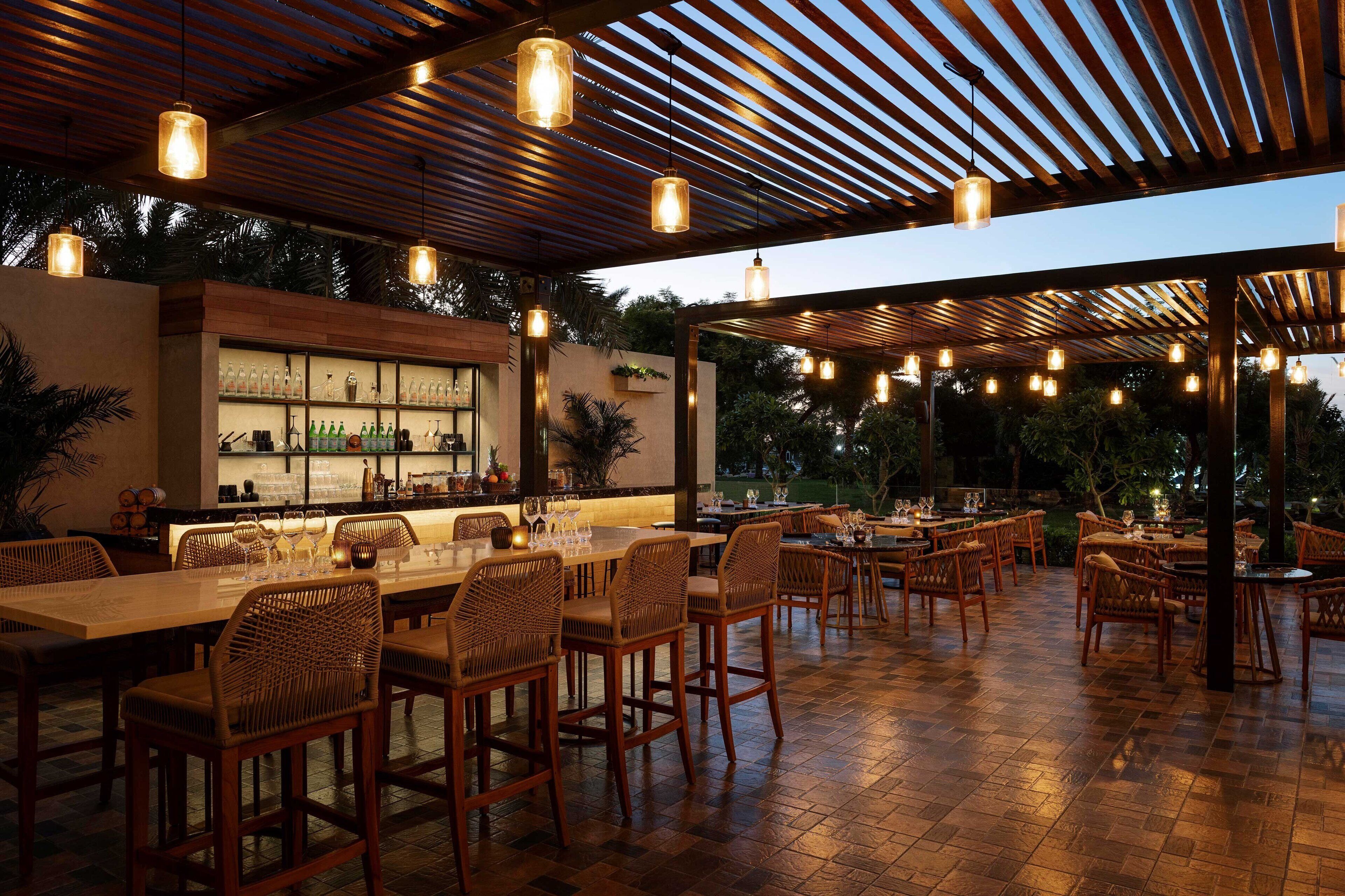 An outdoor dining area with wooden tables and chairs, a bar with shelves of bottles, and hanging pendant lights.