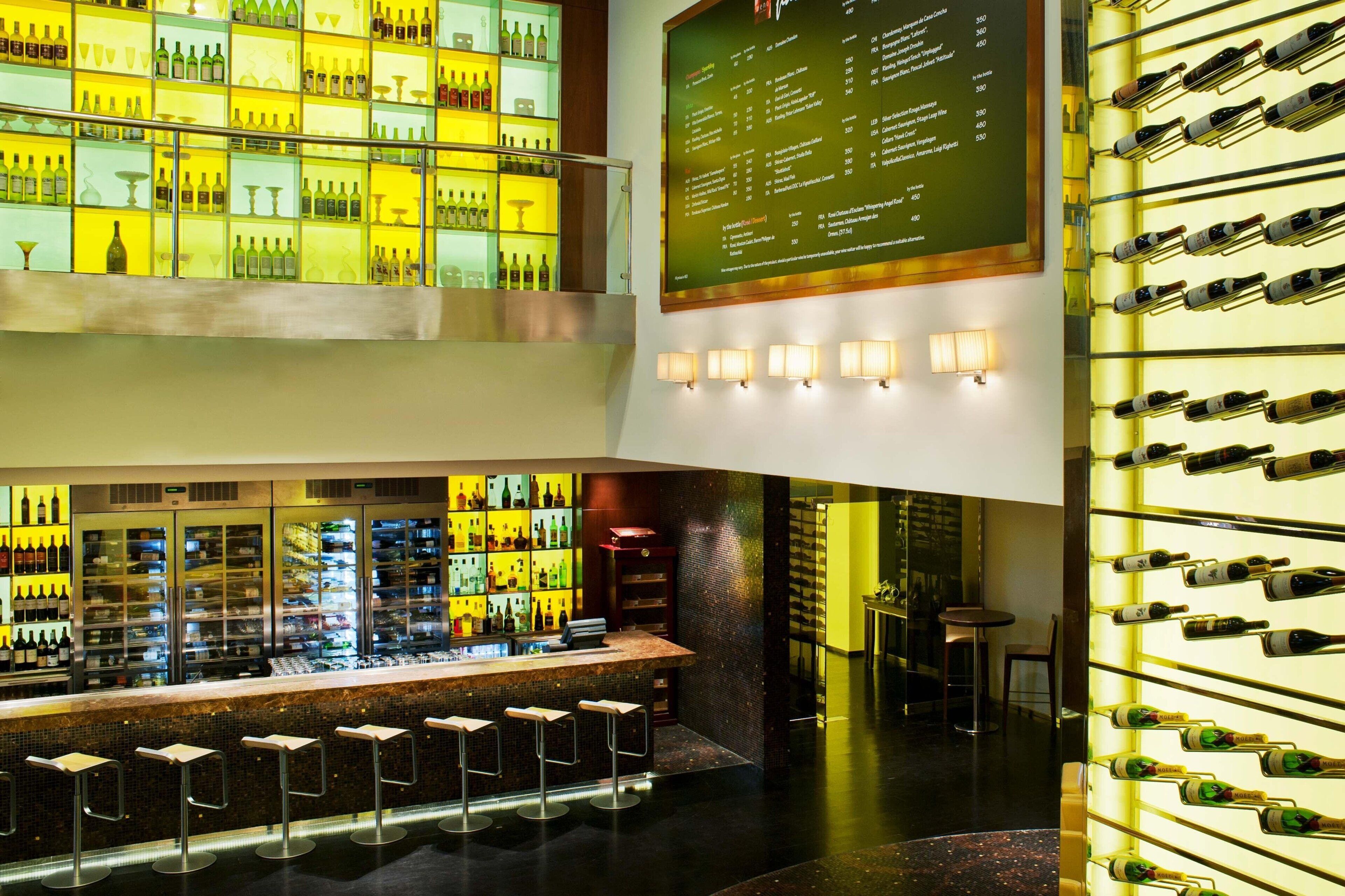 Zona de bar con varias estanterías llenas de botellas, un tablero de menú y taburetes.