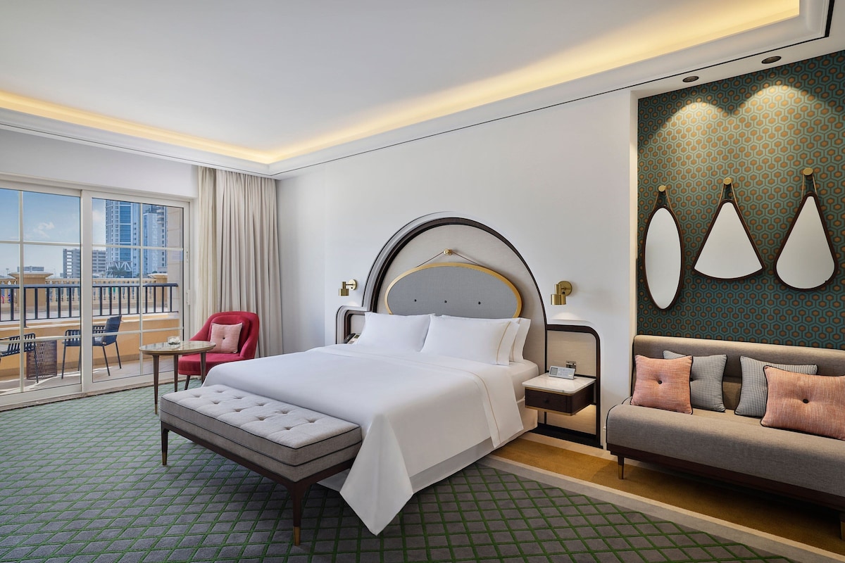 Une chambre d’hôtel avec un grand lit, un canapé, une petite table et une chaise. Il y a une fenêtre donnant sur les bâtiments à l’extérieur.