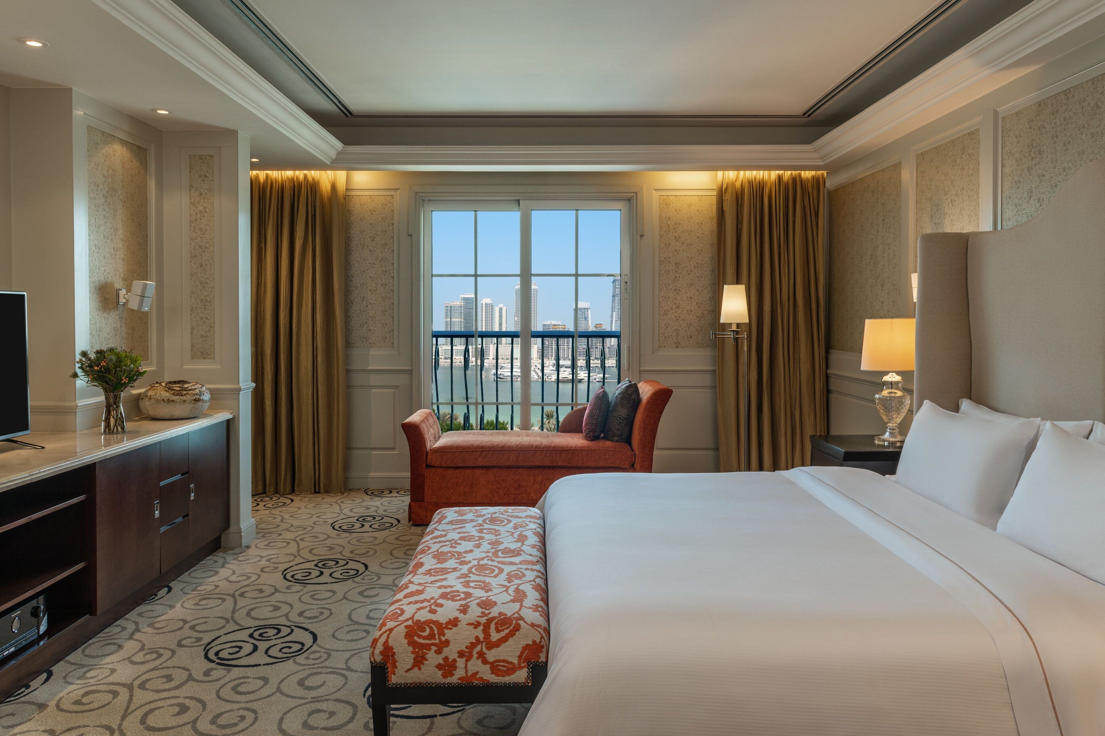 Ein Hotelzimmer mit einem großen Bett, einem Schreibtisch mit Fernseher, einer Sitzbank und Blick auf die Stadt durch ein blickdichtes Fenster.