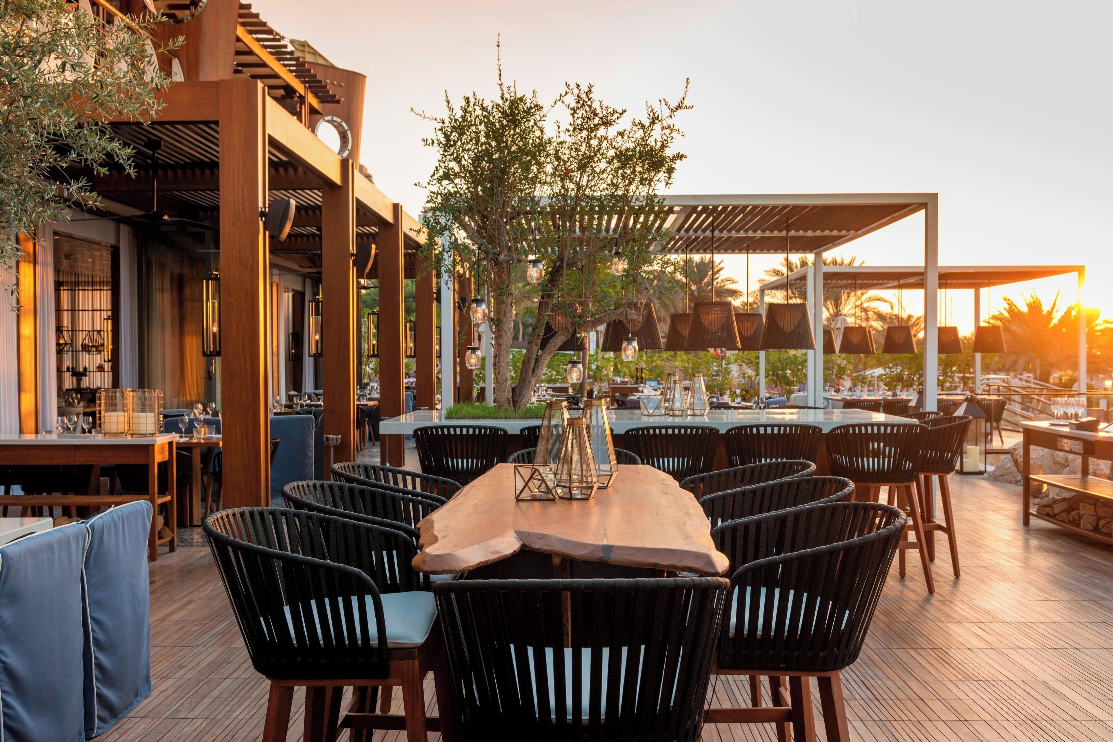 Un área de comedor al aire libre con varias mesas y sillas, rodeada de pérgolas de madera y árboles.