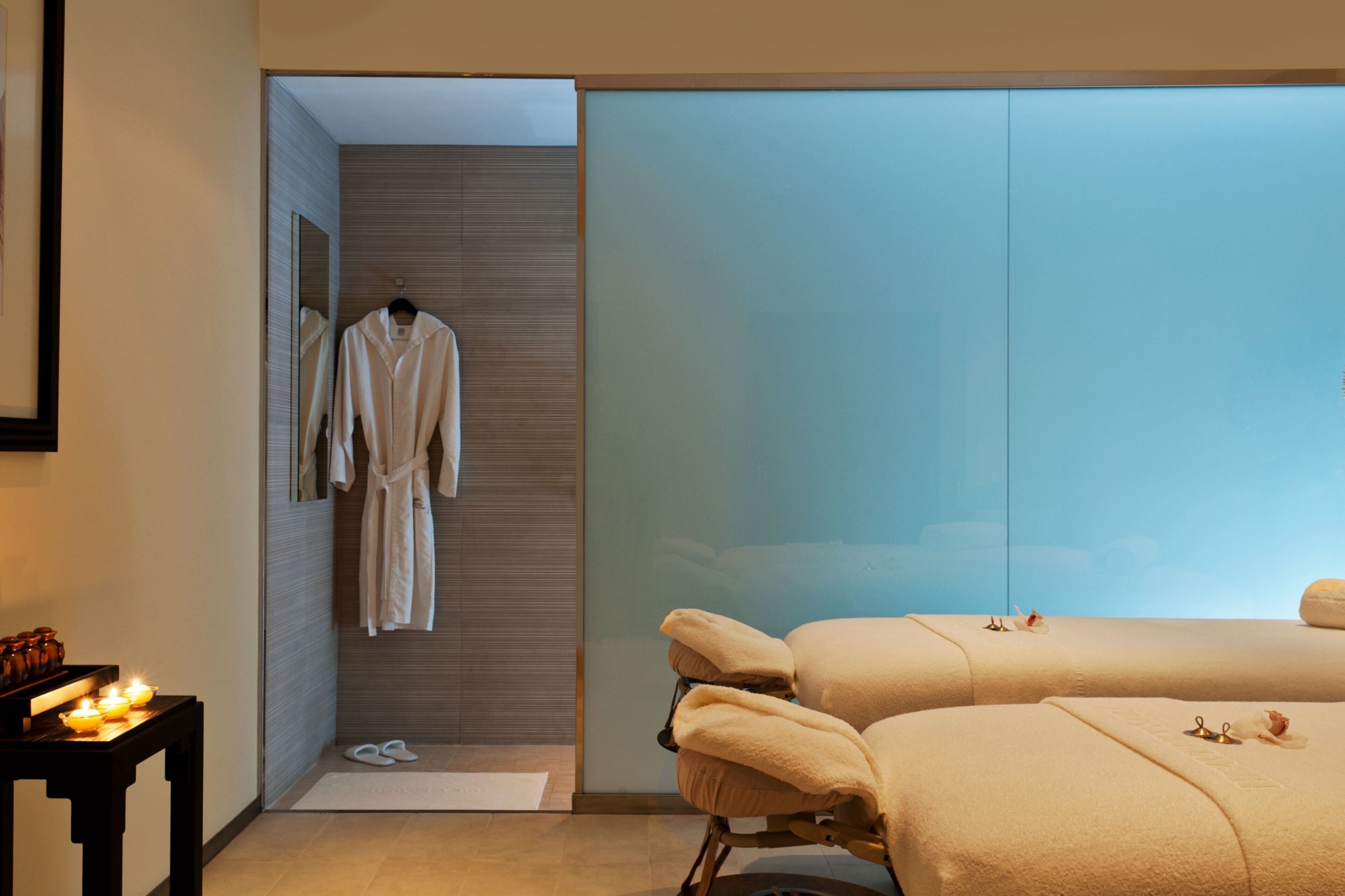 Ein modernes Hotelzimmer mit Glastrennwand, einem an der Wand hängenden Bademantel und zwei Massageliegen.