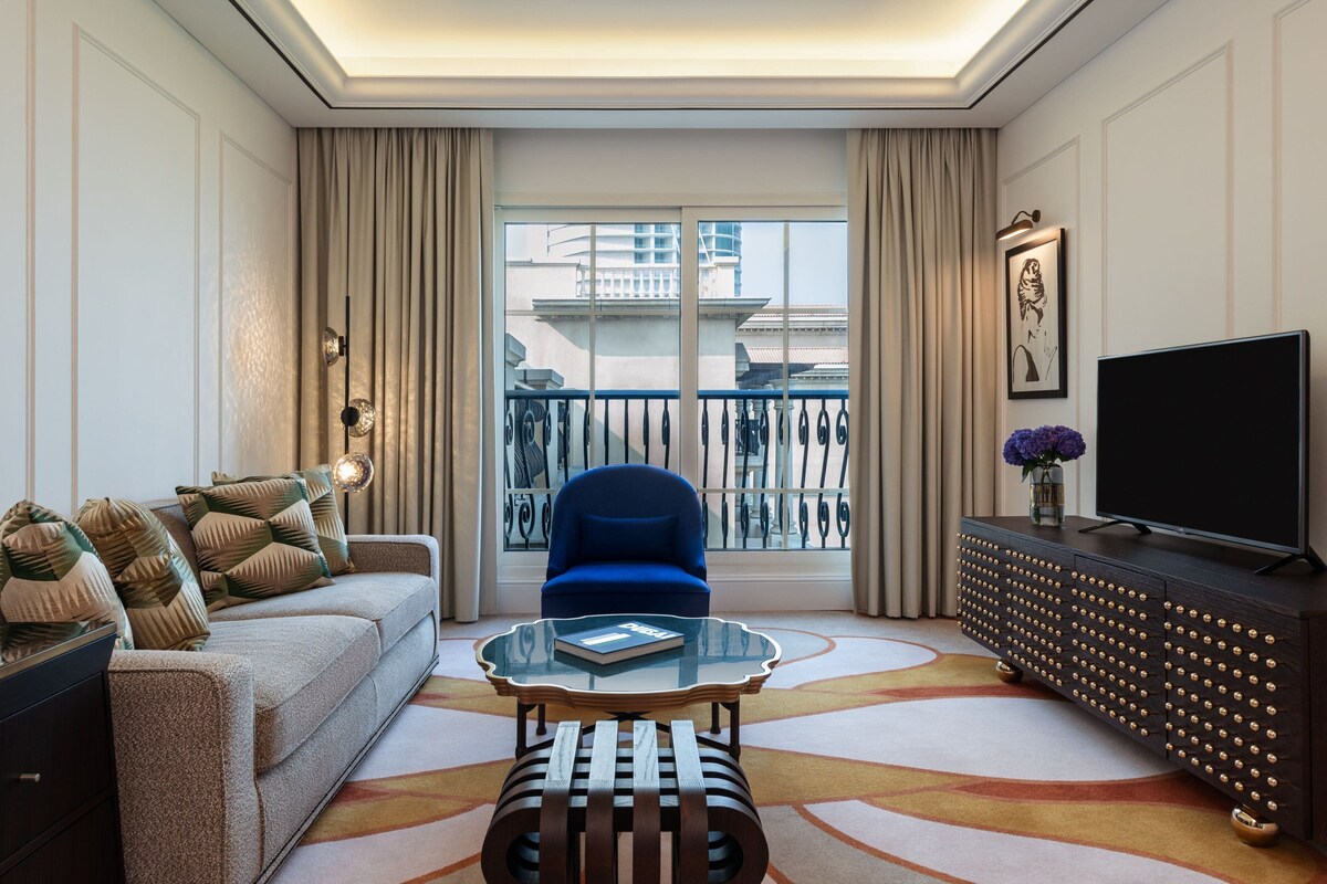Un salon moderne avec un canapé, un fauteuil bleu, une table basse en verre, un téléviseur posé sur un meuble en bois foncé, et une vue sur les immeubles à travers une grande fenêtre.