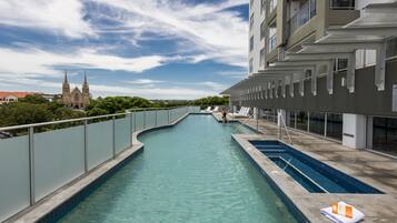 A swimming pool with a curved design, surrounded by a high fence, and a building with multiple balconies in the background.