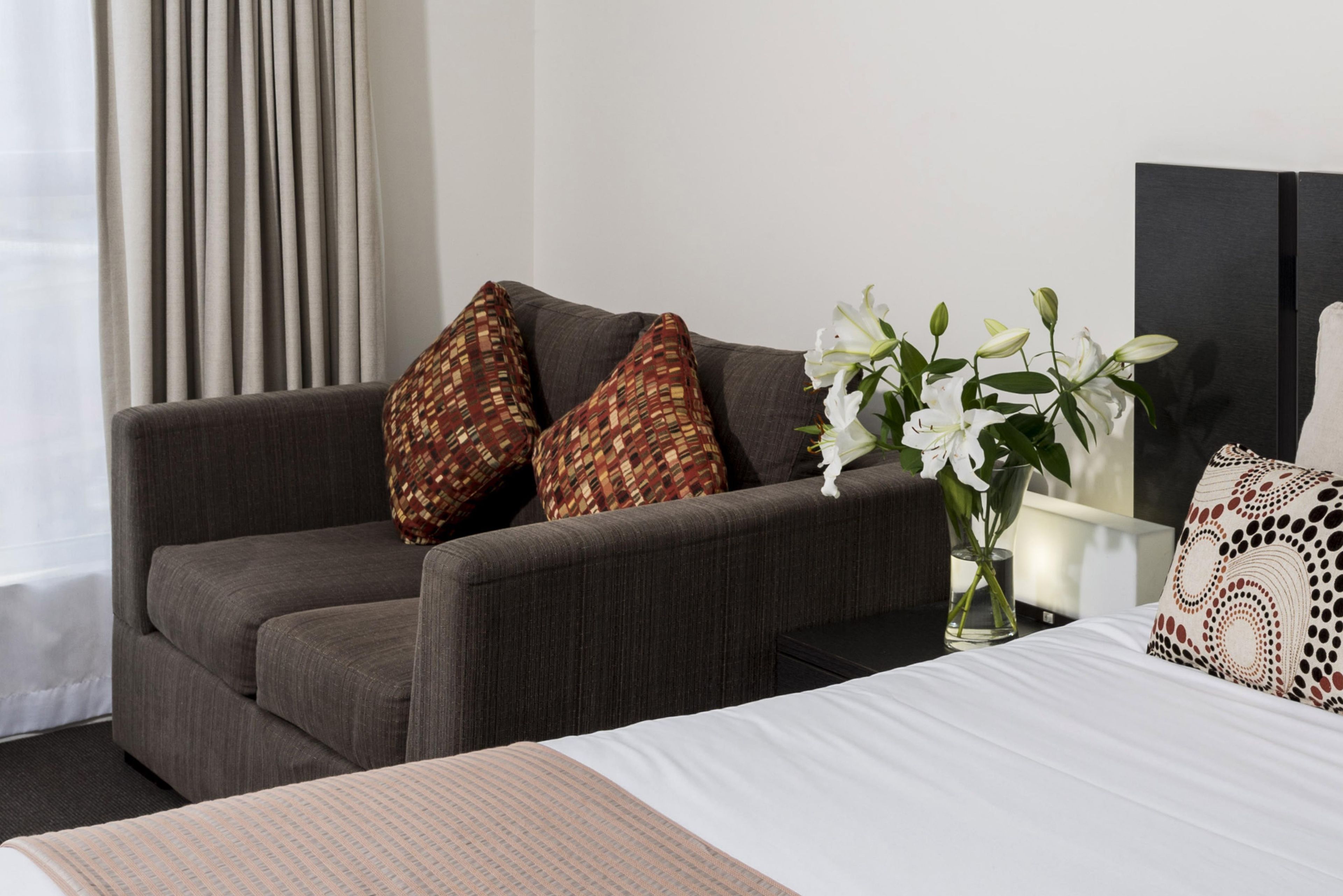 A hotel room with a grey sofa, patterned pillows, a white bed, and a vase with white flowers.