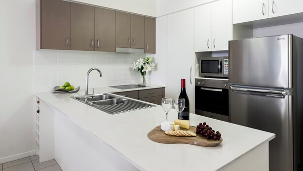 A modern kitchen with white countertops, stainless steel appliances, and a wooden cutting board with cheese and grapes.