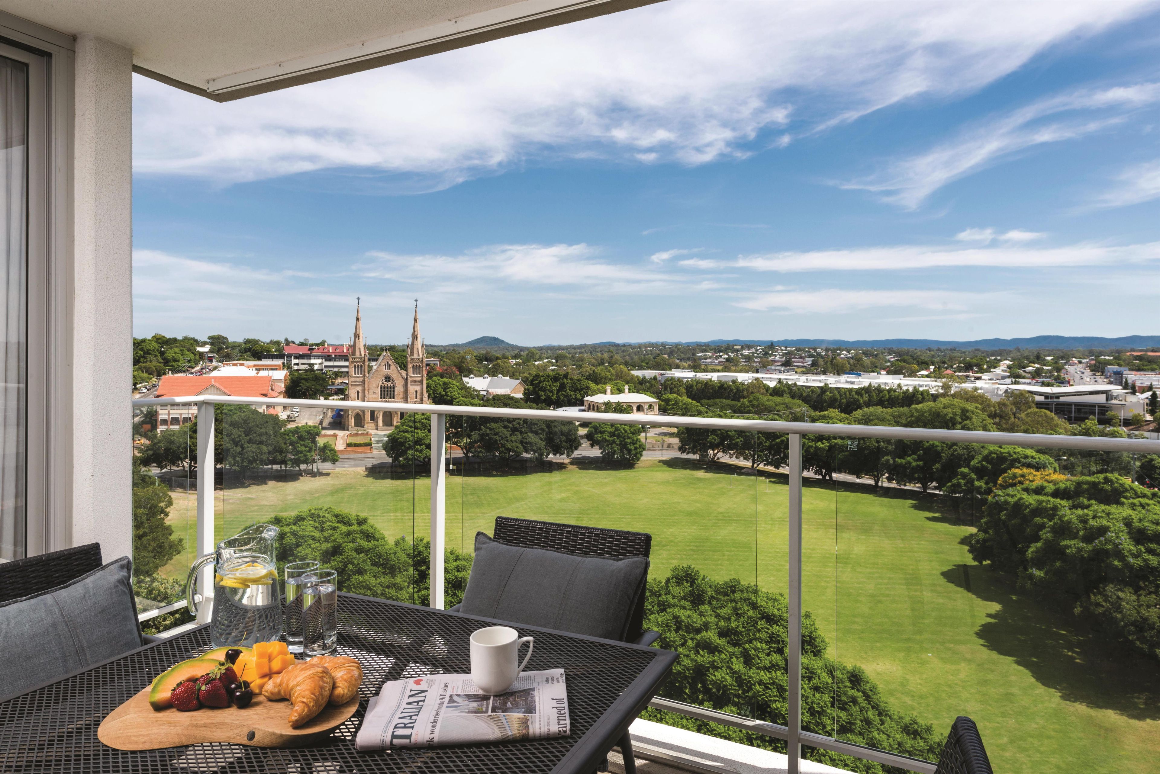 Un balcon avec une table dressée pour le petit-déjeuner, offrant une vue sur le paysage urbain, avec une église et un parc verdoyant.