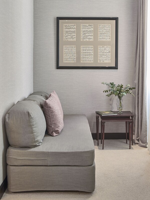 A neutral-toned room with a grey sofa, two cushions, a small wooden side table with a book and vase, and a framed sheet music on the wall.