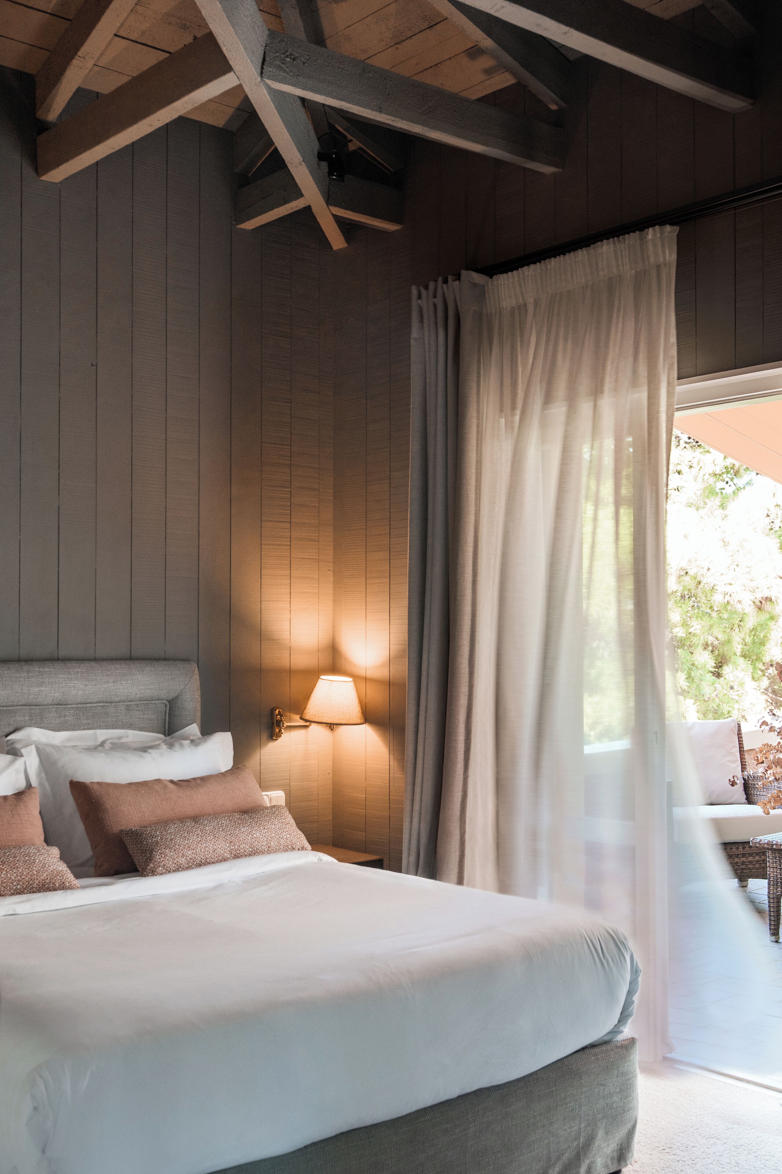 Un dormitorio con una cama grande, una mesita de noche, una lámpara de pared y una ventana con cortinas.