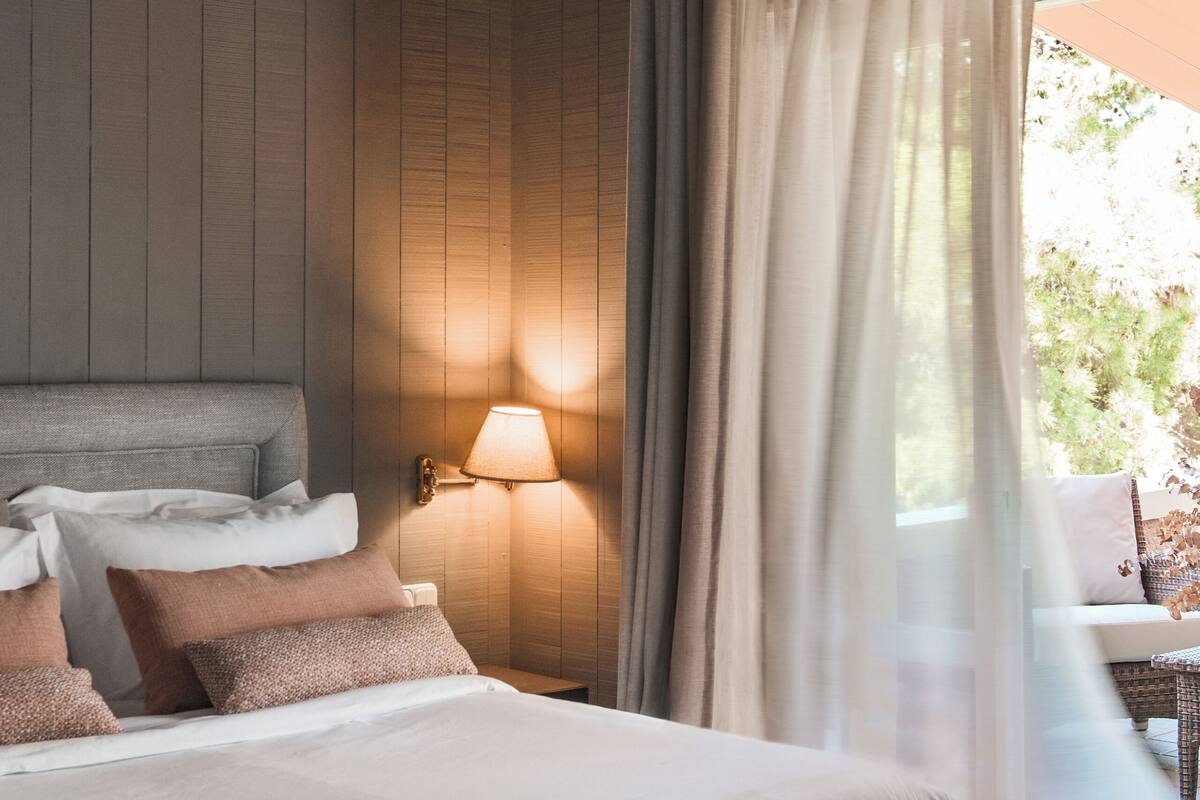 Un dormitorio con una cama grande, una mesita de noche, una lámpara de pared y una ventana con cortinas.