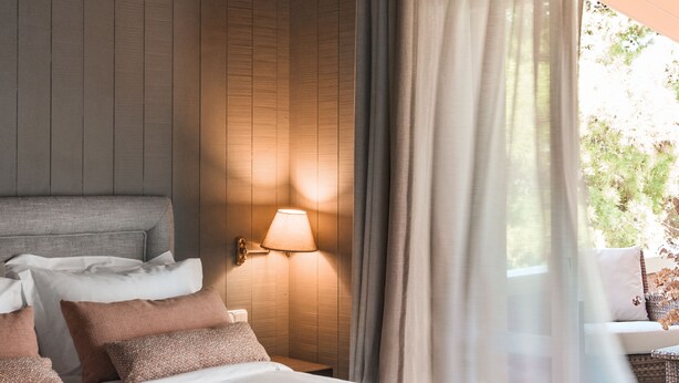 A bedroom with a large bed, a nightstand, a wall-mounted lamp, and a window with curtains.