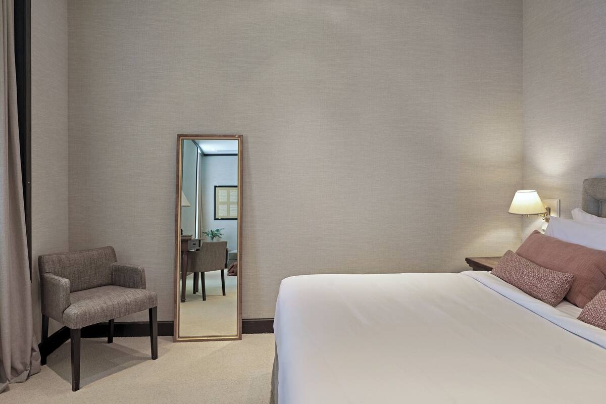 Una habitación de hotel con cama, sillón, mesita de noche, lámpara y espejo.