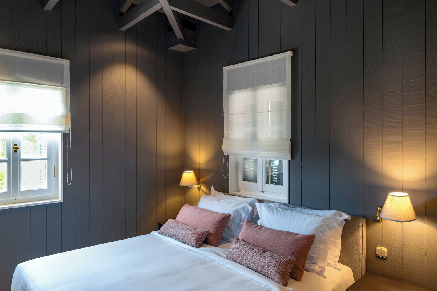 Un dormitorio con cama, dos lámparas de noche, una ventana con persianas y techo con vigas a la vista.