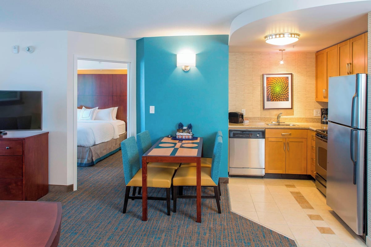 A hotel room with a bed, a dining table with chairs, a TV, a kitchen area with a refrigerator, and a bathroom.