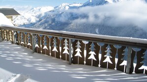 Un balcón cubierto de nieve con baranda de madera y diseños de árboles, con vista a un paisaje montañoso de picos nevados y un valle boscoso.
