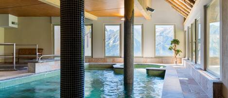 An indoor swimming pool with tiled walls and a wooden ceiling, surrounded by windows with mountain views.