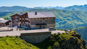 Un edificio de varios pisos con base de piedra, un área de estacionamiento y un paisaje montañoso al fondo.