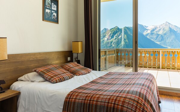 A hotel room with a large bed, a wooden headboard, a nightstand, a lamp, and a view of mountains through a balcony.