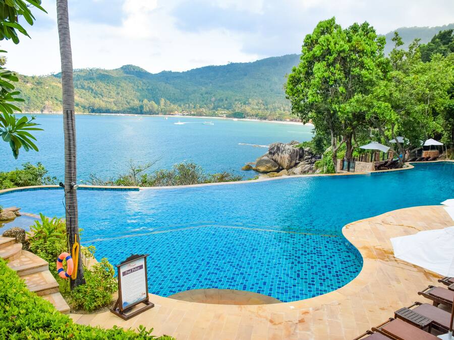 Ein Pool mit geschwungener Kante, umgeben von üppiger Vegetation und Blick auf ein ruhiges Meer und ferne Berge.