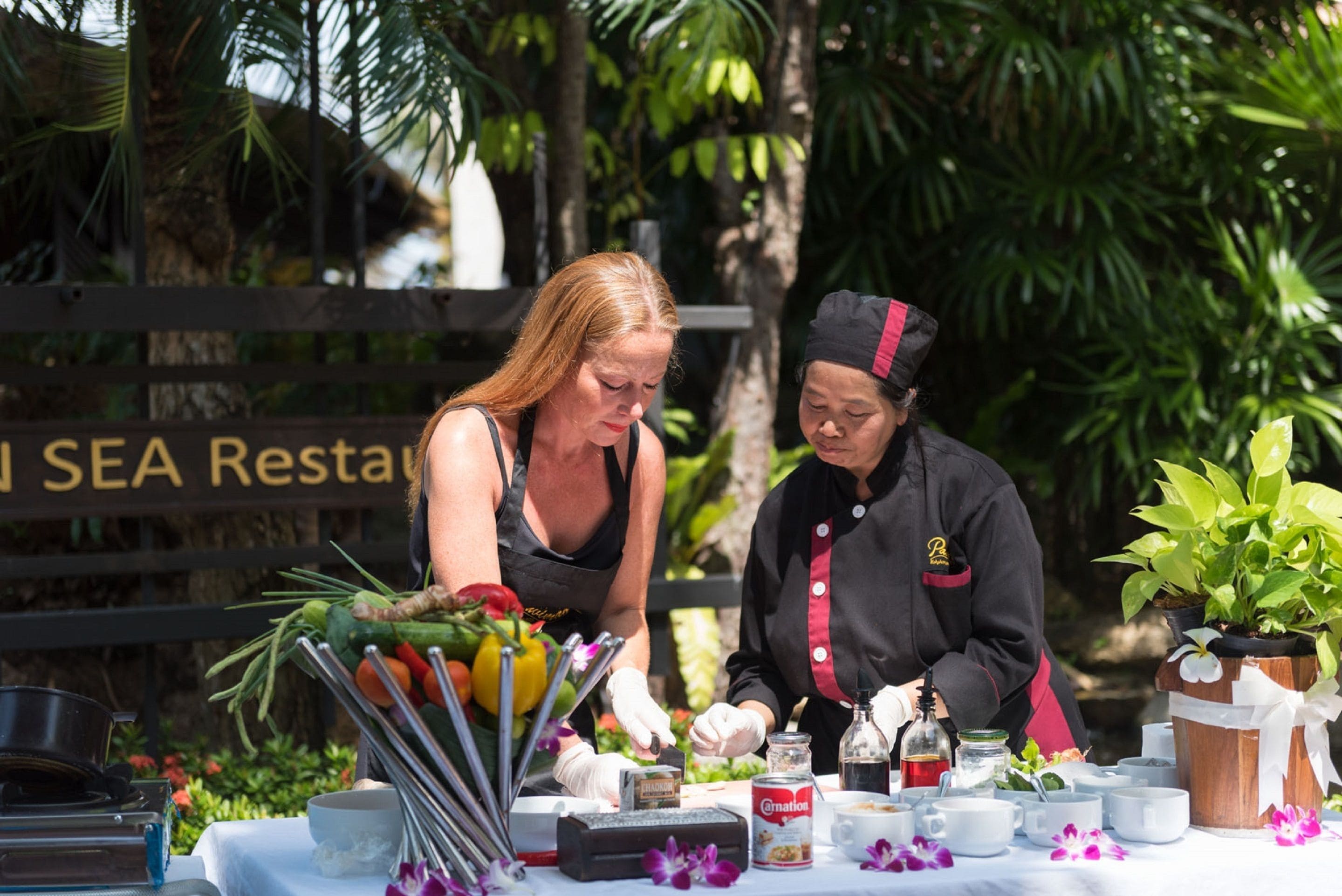 Two individuals preparing food outdoors with a table set with various cooking utensils and ingredients.