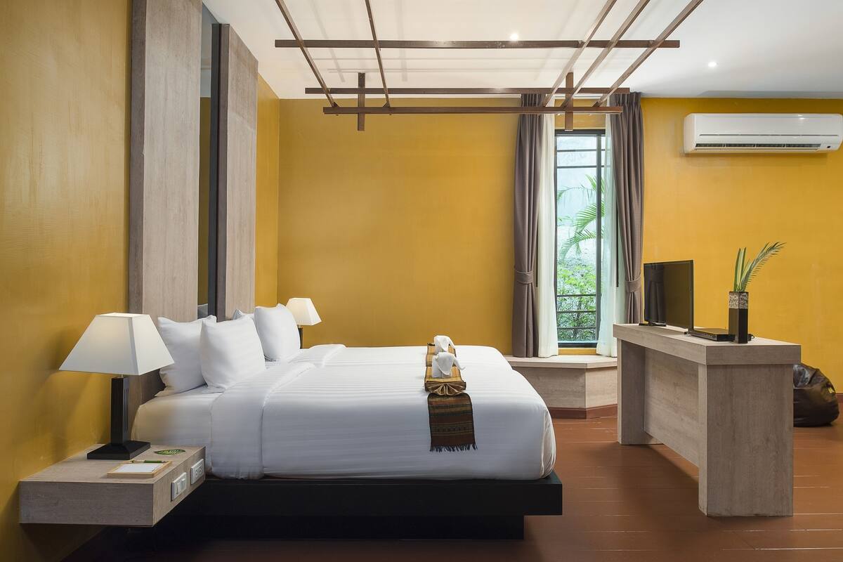 A modern hotel room with a large bed, a bedside table with a lamp, a small desk, and a view of greenery through a window.