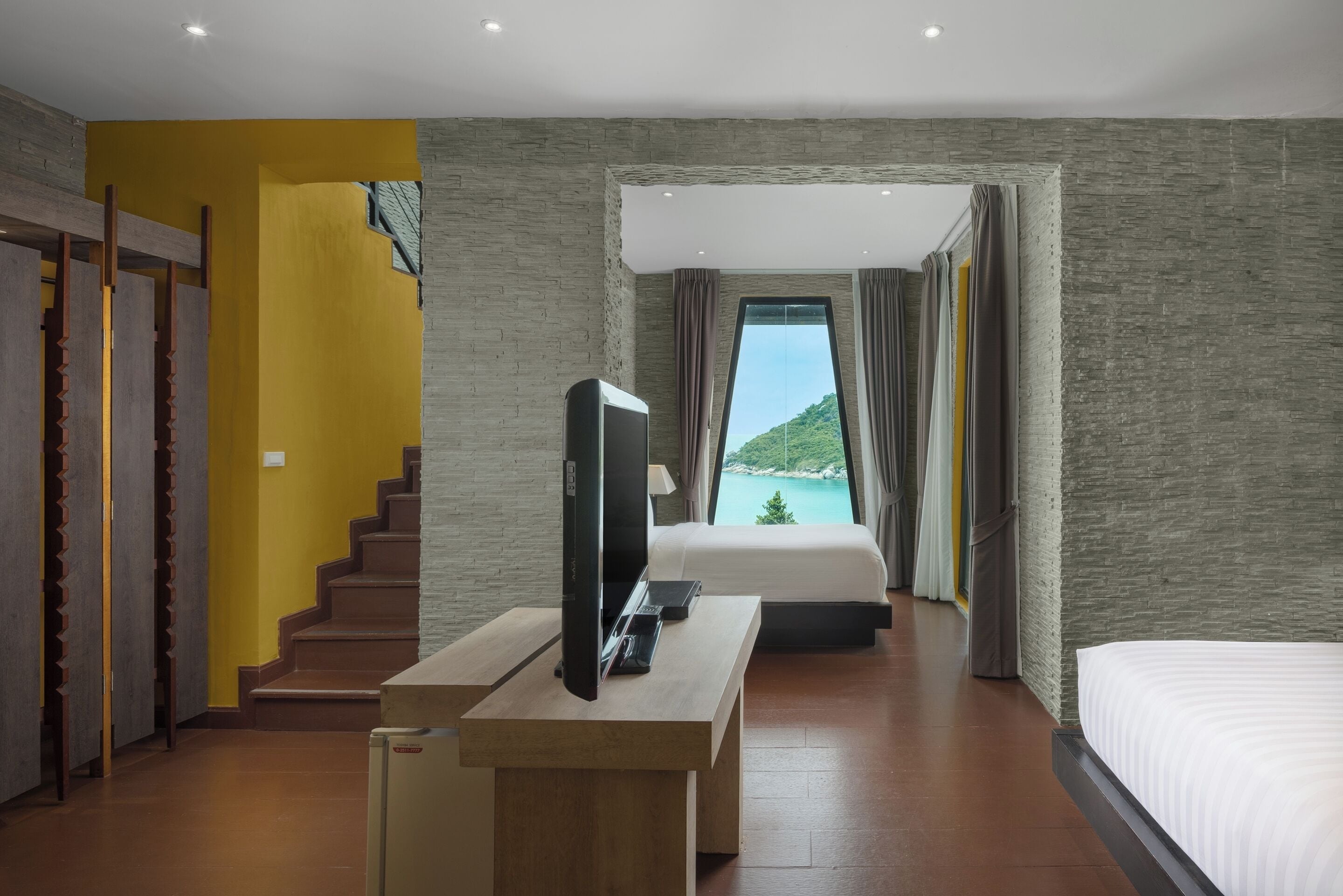 A hotel room with a bed, a TV on a wooden table, a staircase, and a view of a mountain and sea.