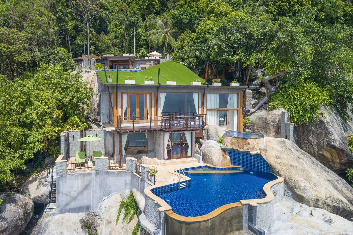 A modern villa with a large swimming pool, a balcony, and lush greenery surrounding it.