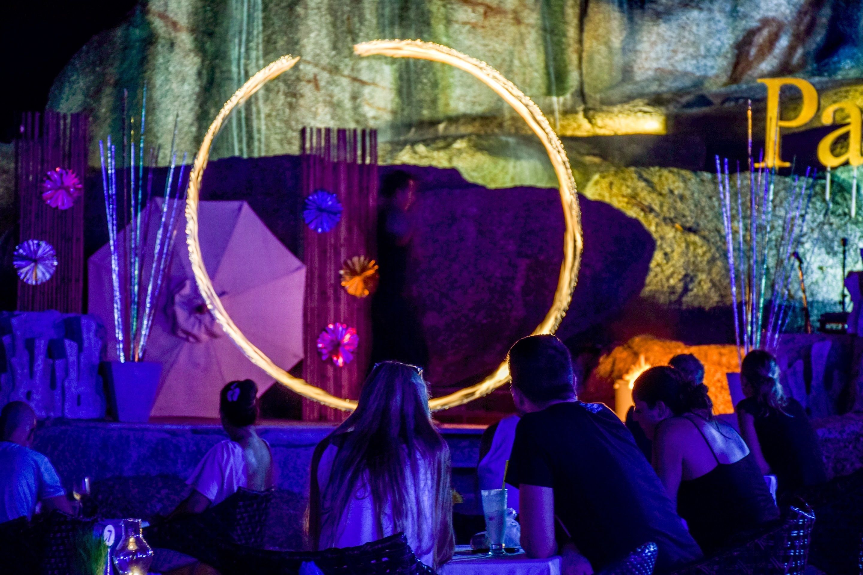 A dimly lit room with a circular light installation, tables with drinks, and a stone wall in the background.