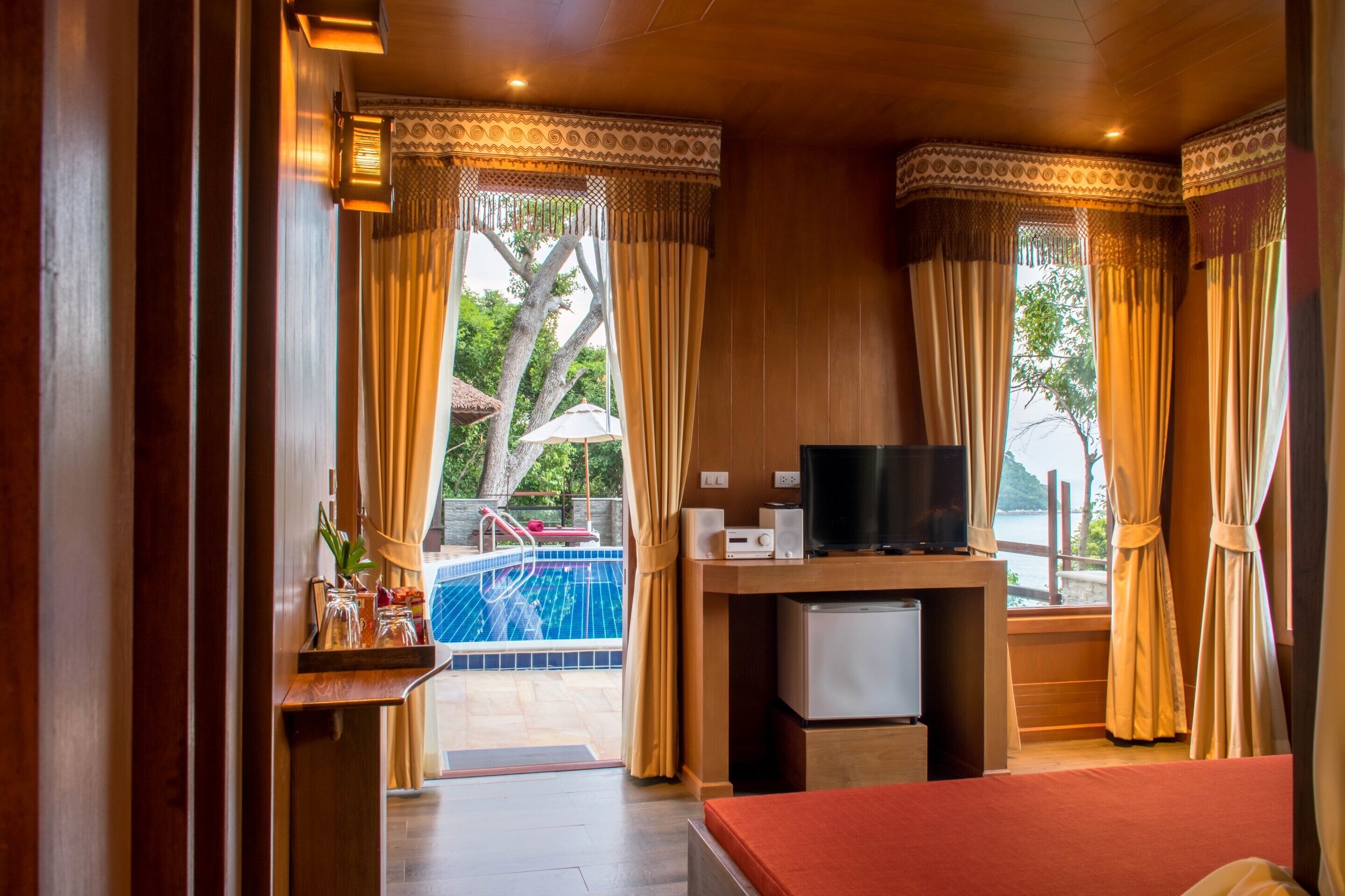 A hotel room with a wooden interior, a television, a small refrigerator, and a view of a swimming pool.