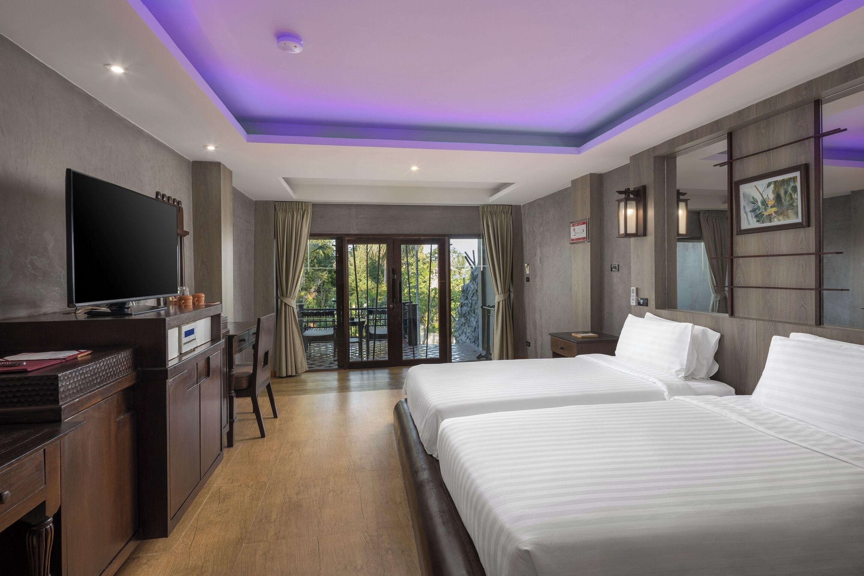 Habitación de hotel con dos camas, televisión, escritorio y balcón con vista a zonas verdes.